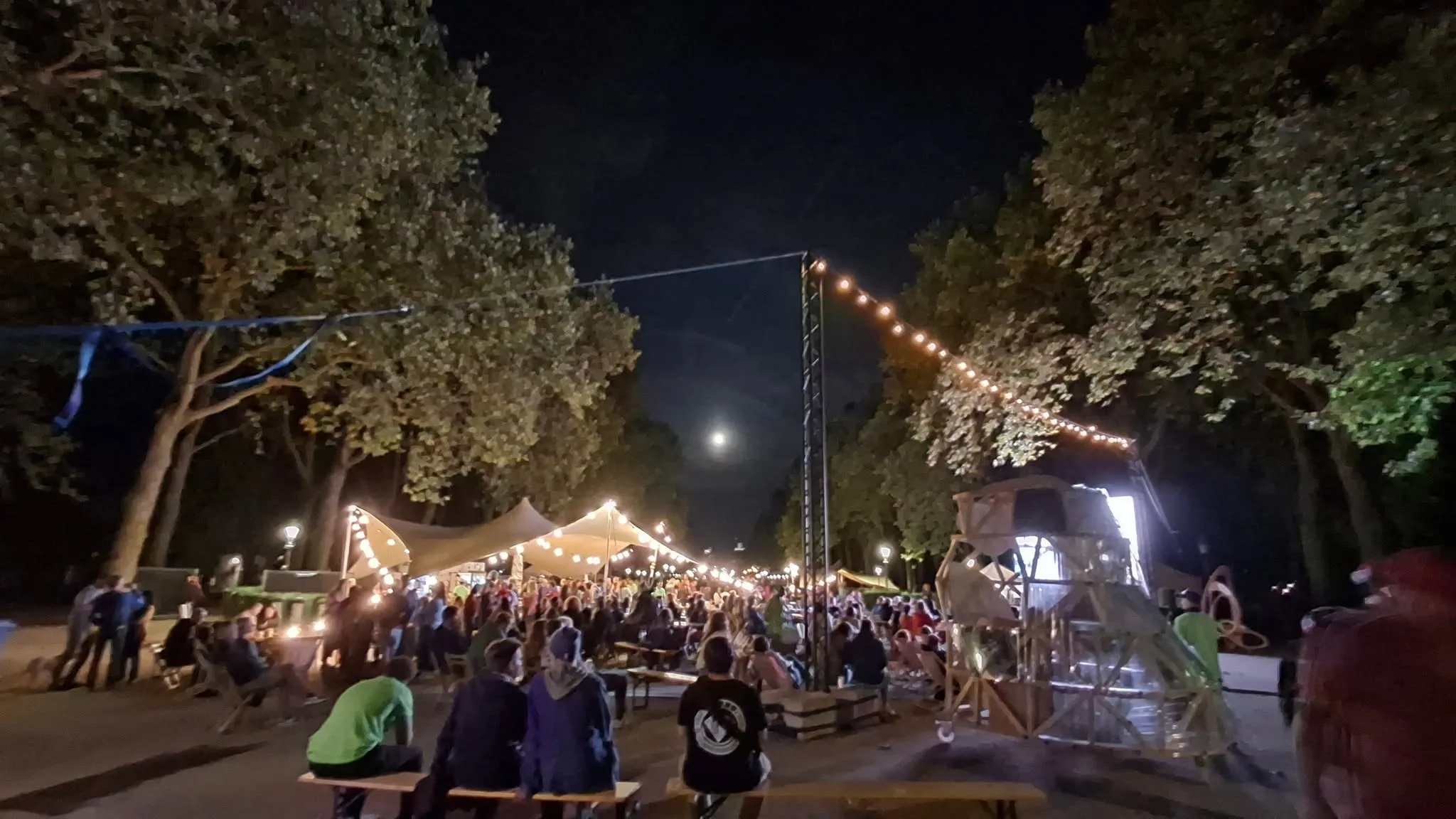 Le site du festival la nuit. Des guirlandes lumineuses entre les arbres, des gens aux tables de pique-nique, la lune au-dessus du parc.