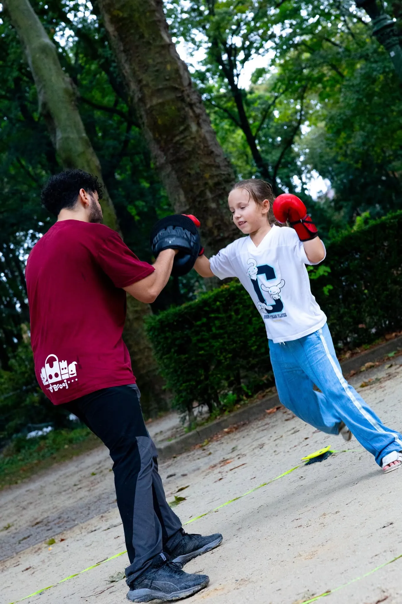 Une fille avec des gants de boxe rouges frappe les pattes d'ours d'un animateur, sur une allée du parc.