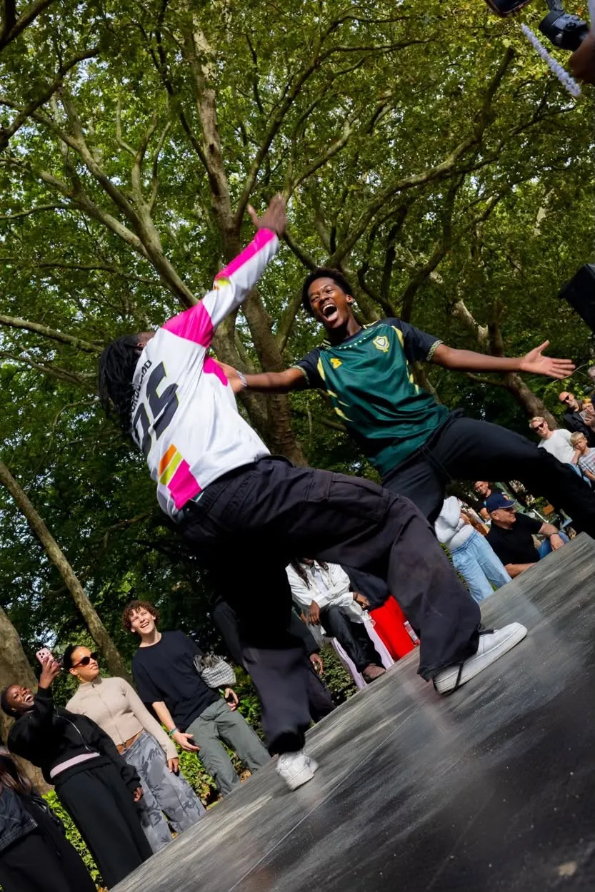 Deux breakdanceurs s'affrontent sur une scène entre les arbres, le public les encourage.