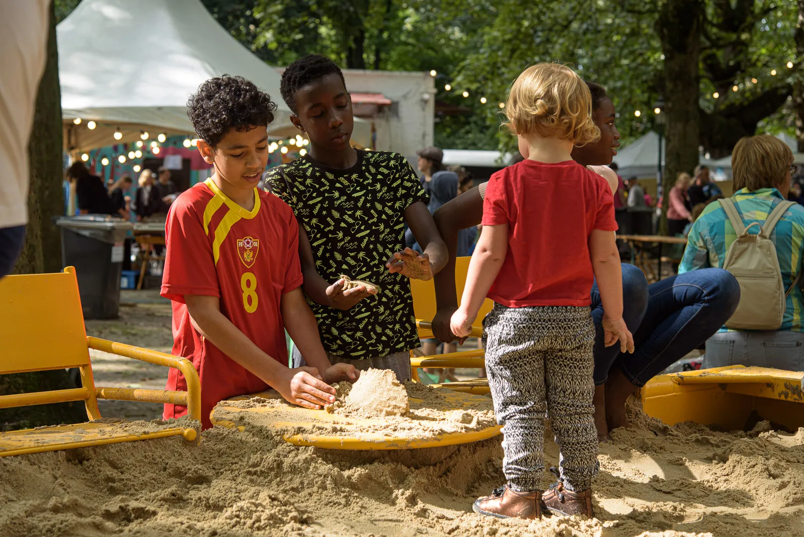 Des enfants d'âges différents jouent ensemble dans un bac à sable à Plazey, avec les stands du festival et des guirlandes en arrière-plan.