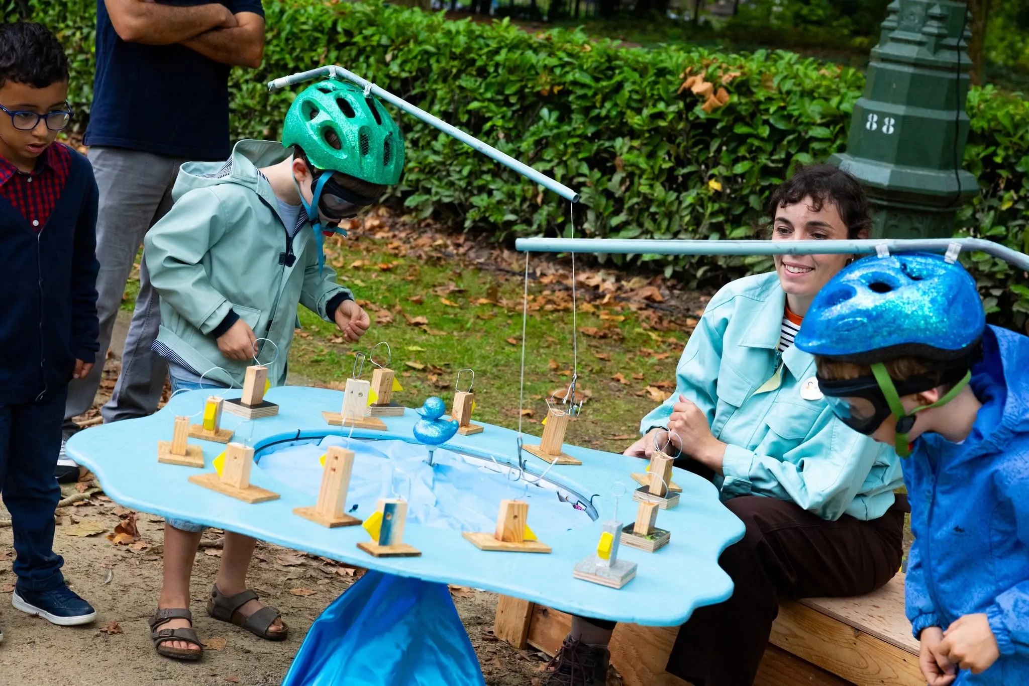 Des enfants avec des casques de vélo jouent autour d'une table à eau bleue avec des petites cannes en bois.