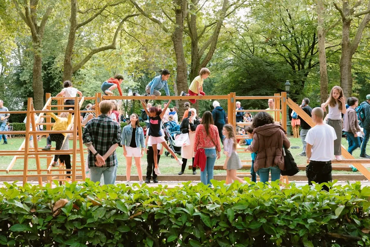 Des enfants grimpent sur une structure en bois dans le parc, les parents regardent depuis la pelouse.
