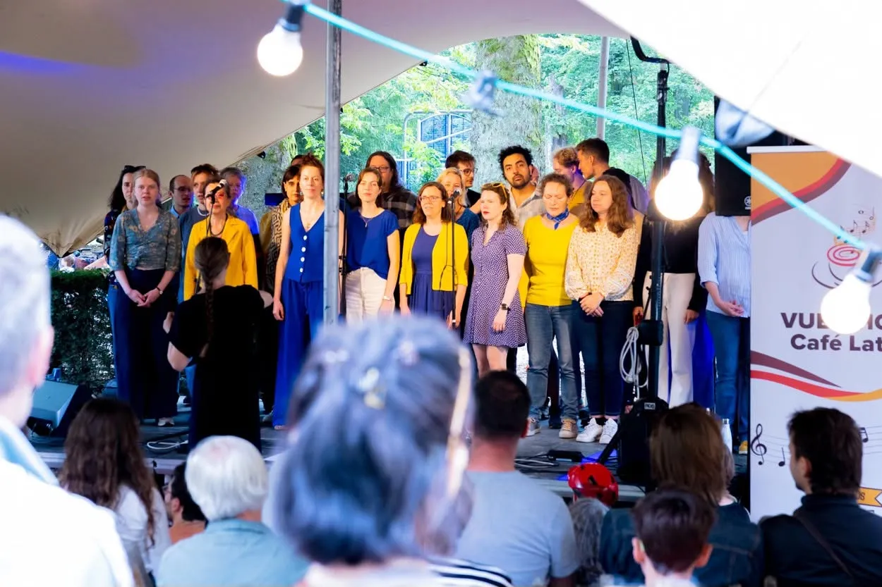 Une chorale en bleu et jaune chante sous la tente, avec une bannière du Café Latino en arrière-plan.
