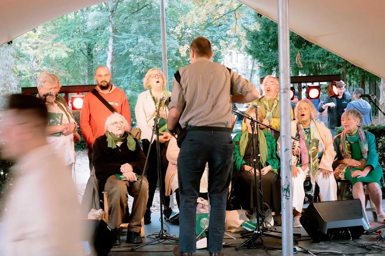 Une chorale mêlant jeunes et moins jeunes chante sous une tente dans le parc, dirigée par un chef de chœur.
