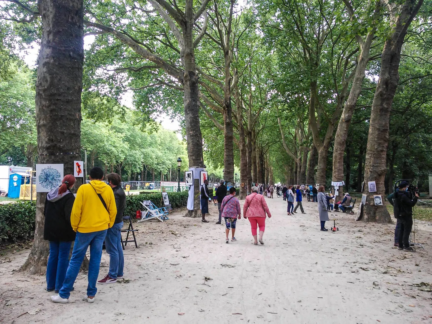 Allée d'arbres du Parc Elisabeth avec des œuvres d'art accrochées aux branches, des visiteurs marchent en dessous.