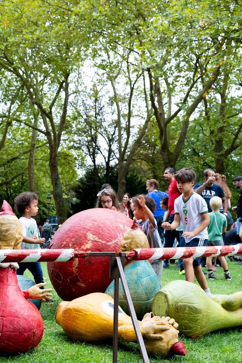 D'énormes fruits en papier mâché sont allongés dans l'herbe, des enfants les regardent avec curiosité.