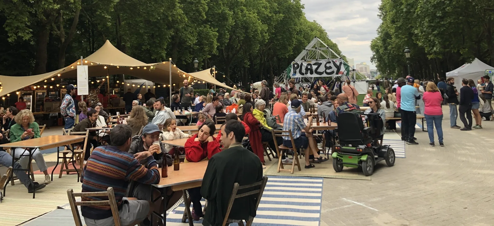 Le site du festival Plazey au parc Élisabeth : tables de pique-nique, guirlandes et le panneau Plazey entre les arbres.