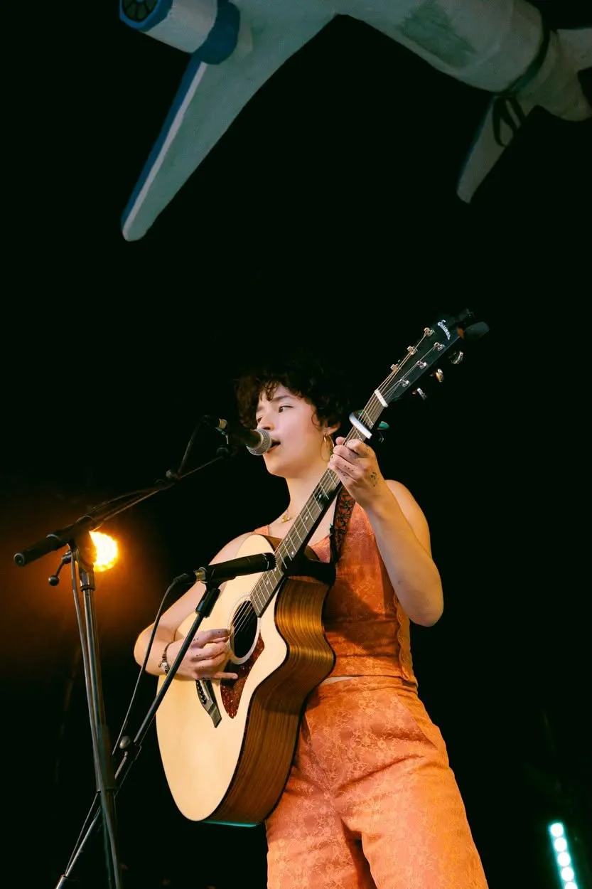 Une chanteuse en tenue orange joue de la guitare acoustique, sous l'aile de l'avion.